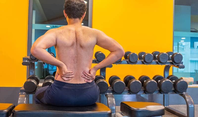 Shirtless man holding lower back pain in gym bench.